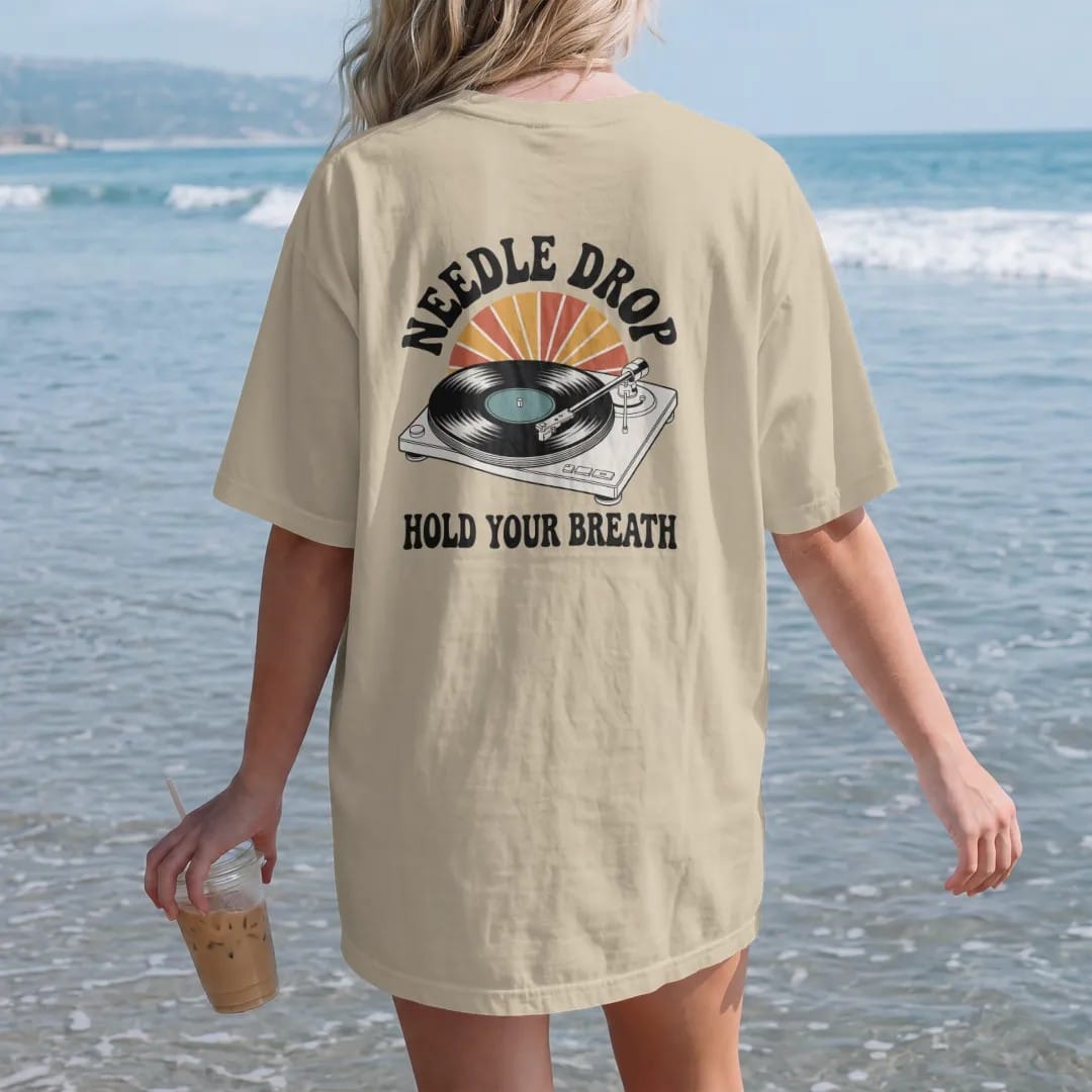 Woman on the beach in a Sandstone tee, back view: a turntable tonearm lowering onto a spinning record, with "NEEDLE DROP" and "HOLD YOUR BREATH" in chunky 70s lettering and small 70s caps, printed in faded, cracked cream, mustard, burnt orange and…