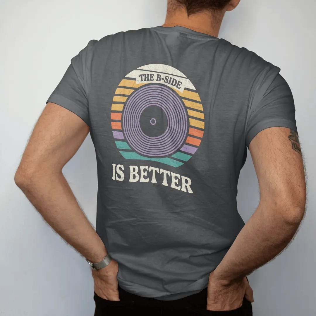 Man wearing a Graphite tee, seen from the back: a large record center label surrounded by groove rings, with "THE B-SIDE" and "IS BETTER" in chunky 70s lettering and small 70s caps, printed in faded, cracked cream, mustard, burnt orange and faded…