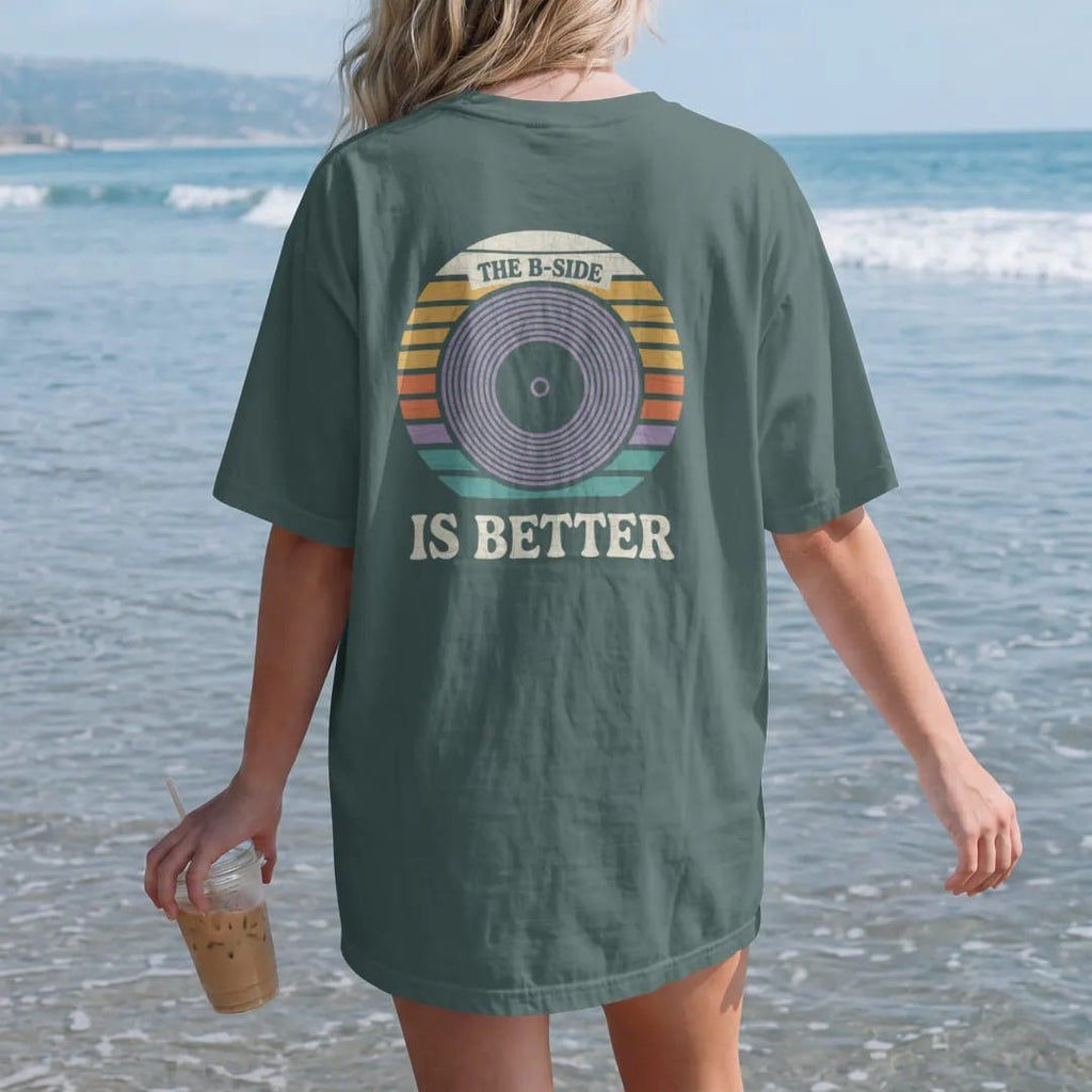 Woman on the beach in a Blue Spruce tee, back view: a large record center label surrounded by groove rings, with "THE B-SIDE" and "IS BETTER" in chunky 70s lettering and small 70s caps, printed in faded, cracked cream, mustard, burnt orange and…