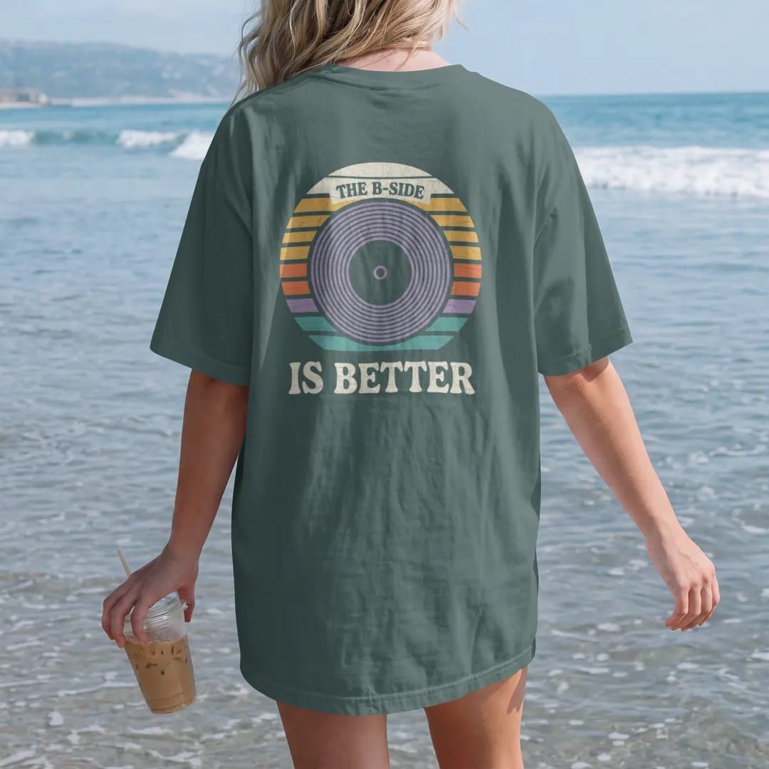 Woman on the beach in a Blue Spruce tee, back view: a large record center label surrounded by groove rings, with "THE B-SIDE" and "IS BETTER" in chunky 70s lettering and small 70s caps, printed in faded, cracked cream, mustard, burnt orange and…