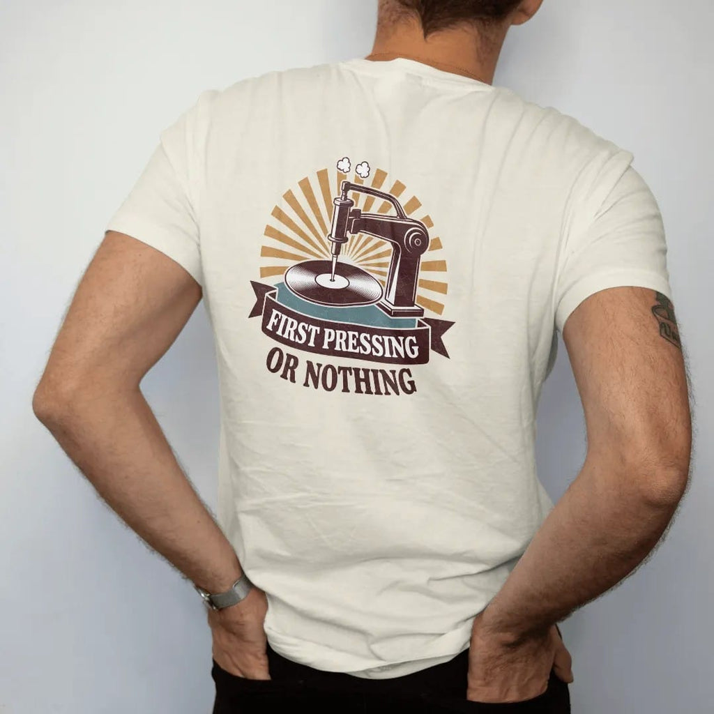 Man wearing a Ivory tee, seen from the back: a vintage record press stamping a fresh disc, with "FIRST PRESSING" and "OR NOTHING" in chunky 70s lettering and small 70s caps, printed in faded, cracked brown, burnt orange, faded teal and mustard over…