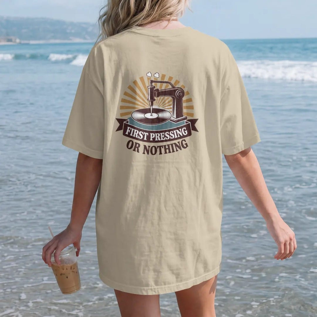 Woman on the beach in a Sandstone tee, back view: a vintage record press stamping a fresh disc, with "FIRST PRESSING" and "OR NOTHING" in chunky 70s lettering and small 70s caps, printed in faded, cracked brown, burnt orange, faded teal and mustard…