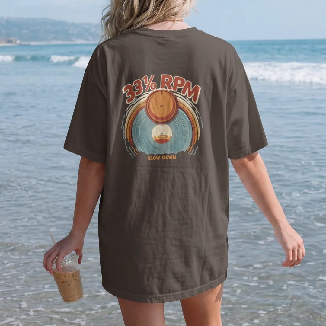 Woman on the beach in a Espresso tee, back view: a turntable speed knob with a sun setting into a spinning record, with "33⅓ RPM" and "SLOW DOWN" in chunky 70s lettering and small 70s caps, printed in faded, cracked cream, mustard, burnt orange and…
