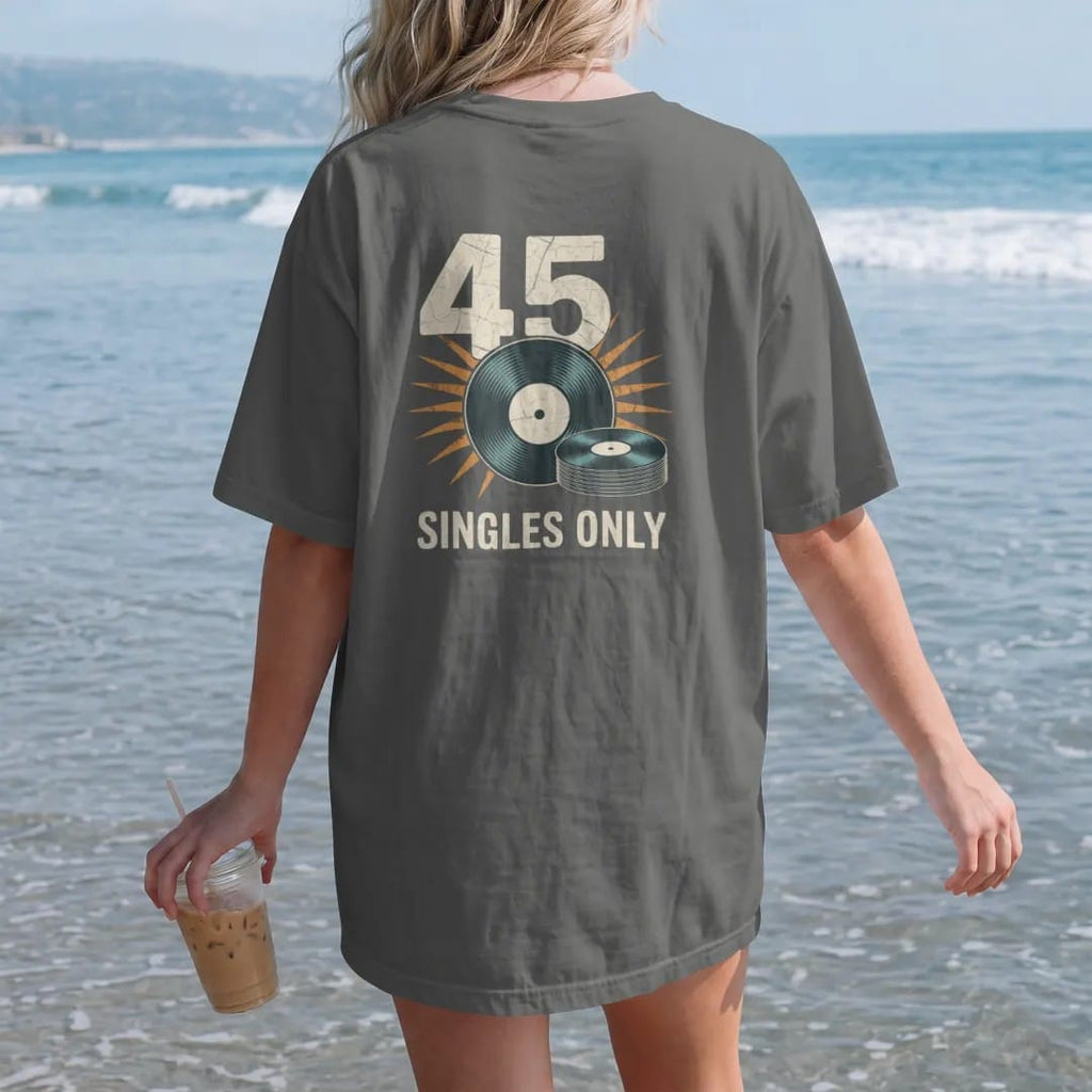 Woman on the beach in a Pepper tee, back view: a large 45 RPM adapter beside a stack of seven-inch records, with "45" and "SINGLES ONLY" in chunky 70s lettering and small 70s caps, printed in faded, cracked cream, mustard, burnt orange and faded…