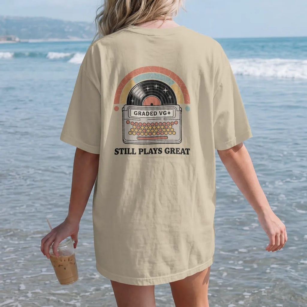 Woman on the beach in a Sandstone tee, back view: a record sleeve with a grading sticker and a few scuffs, with "GRADED VG+" and "STILL PLAYS GREAT" in chunky 70s lettering and small 70s caps, printed in faded, cracked cream, mustard, burnt orange…