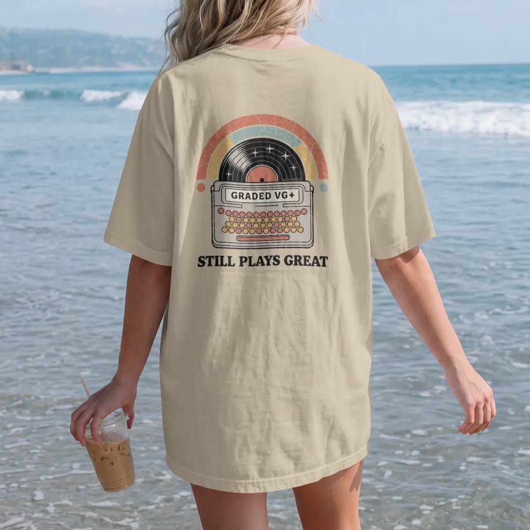 Woman on the beach in a Sandstone tee, back view: a record sleeve with a grading sticker and a few scuffs, with "GRADED VG+" and "STILL PLAYS GREAT" in chunky 70s lettering and small 70s caps, printed in faded, cracked cream, mustard, burnt orange…