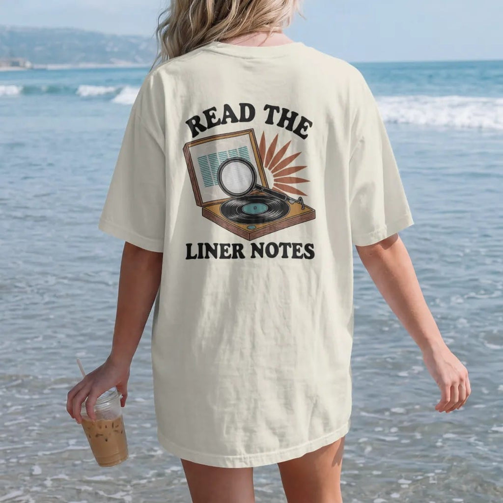 Woman on the beach in a Ivory tee, back view: an open record sleeve under a magnifying glass with reading glasses, with "READ THE" and "LINER NOTES" in chunky 70s lettering and small 70s caps, printed in faded, cracked brown, burnt orange, faded…
