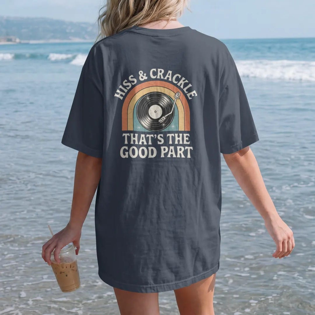 Woman on the beach in a Midnight tee, back view: a spinning record giving off crackle sparks and hiss lines, with "HISS & CRACKLE" and "THAT'S THE GOOD PART" in chunky 70s lettering and small 70s caps, printed in faded, cracked cream, mustard,…