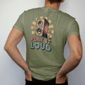 Man wearing a Moss tee, seen from the back: a wooden speaker cabinet sending out sound-wave arcs, with "PLAY IT LOUD" in chunky 70s lettering, printed in faded, cracked cream, mustard, burnt orange and faded teal over a small sunburst.