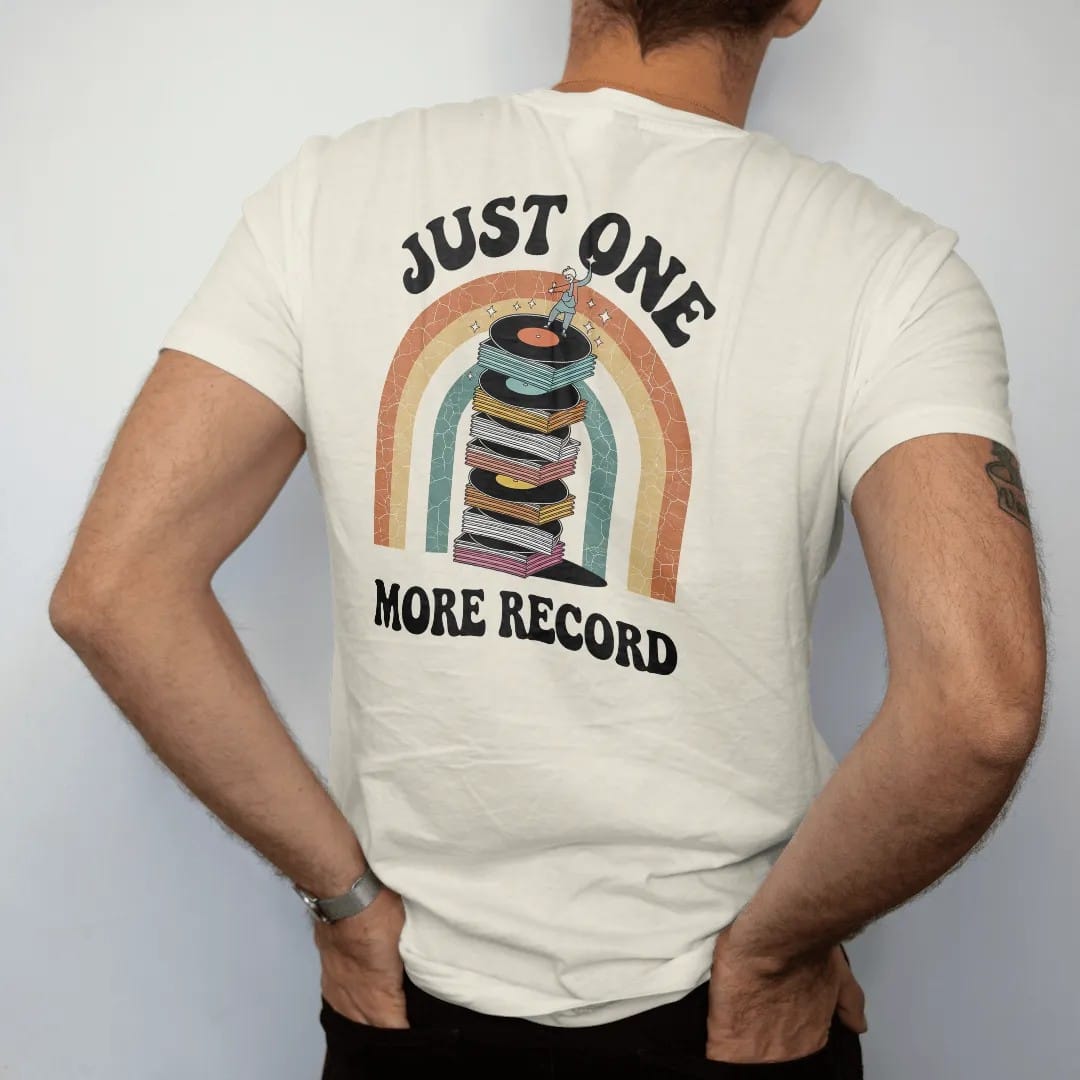 Man wearing a Ivory tee, seen from the back: a teetering stack of records with a tiny person adding one more, with "JUST ONE" and "MORE RECORD" in chunky 70s lettering and small 70s caps, printed in faded, cracked cream, mustard, burnt orange and…