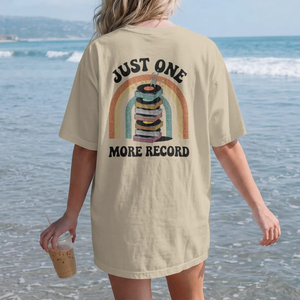 Woman on the beach in a Sandstone tee, back view: a teetering stack of records with a tiny person adding one more, with "JUST ONE" and "MORE RECORD" in chunky 70s lettering and small 70s caps, printed in faded, cracked cream, mustard, burnt orange…