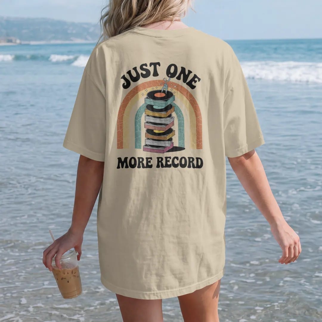 Woman on the beach in a Sandstone tee, back view: a teetering stack of records with a tiny person adding one more, with "JUST ONE" and "MORE RECORD" in chunky 70s lettering and small 70s caps, printed in faded, cracked cream, mustard, burnt orange…