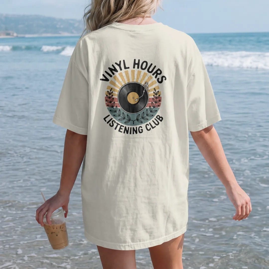Woman on the beach in a Ivory tee, back view: a round club emblem with a record, tonearm and sunburst rays, with "VINYL HOURS" and "LISTENING CLUB" in chunky 70s lettering and small 70s caps, printed in faded, cracked cream, mustard, burnt orange…