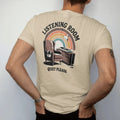 Man wearing a Sandstone tee, seen from the back: a mid-century armchair facing a turntable console with headphones, with "LISTENING ROOM" and "QUIET PLEASE" in chunky 70s lettering and small 70s caps, printed in faded, cracked brown, burnt orange,…