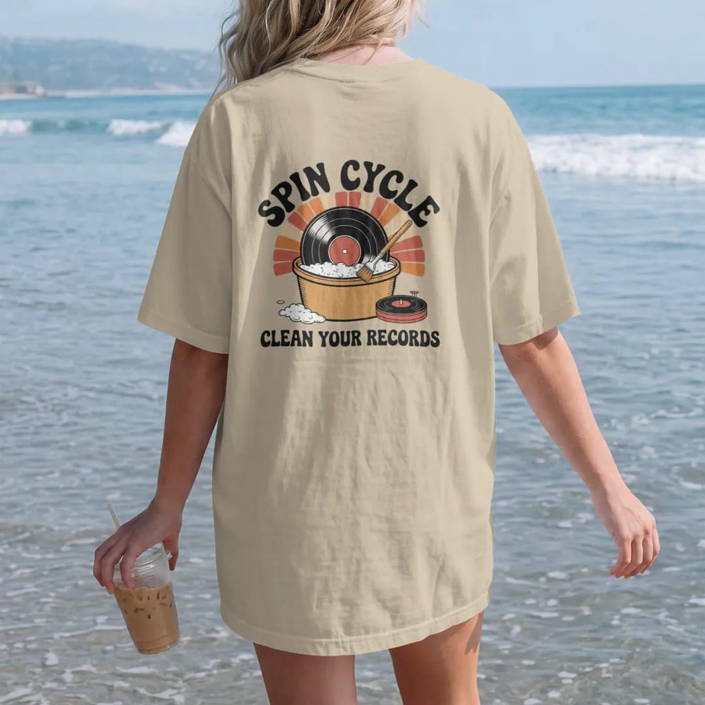 Woman on the beach in a Sandstone tee, back view: a record in a bubble bath beside a cleaning-machine dial, with "SPIN CYCLE" and "CLEAN YOUR RECORDS" in chunky 70s lettering and small 70s caps, printed in faded, cracked cream, mustard, burnt…