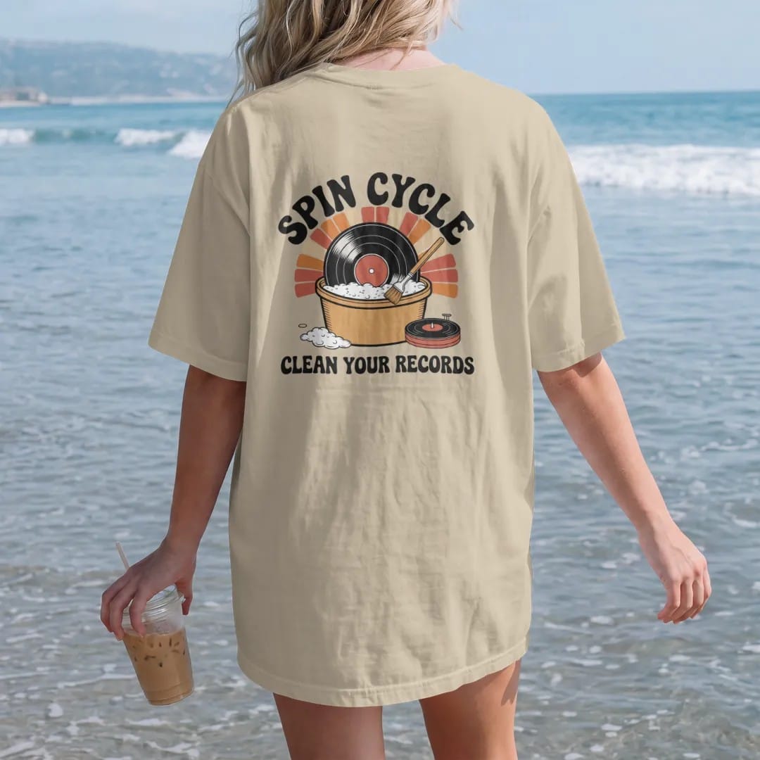 Woman on the beach in a Sandstone tee, back view: a record in a bubble bath beside a cleaning-machine dial, with "SPIN CYCLE" and "CLEAN YOUR RECORDS" in chunky 70s lettering and small 70s caps, printed in faded, cracked cream, mustard, burnt…