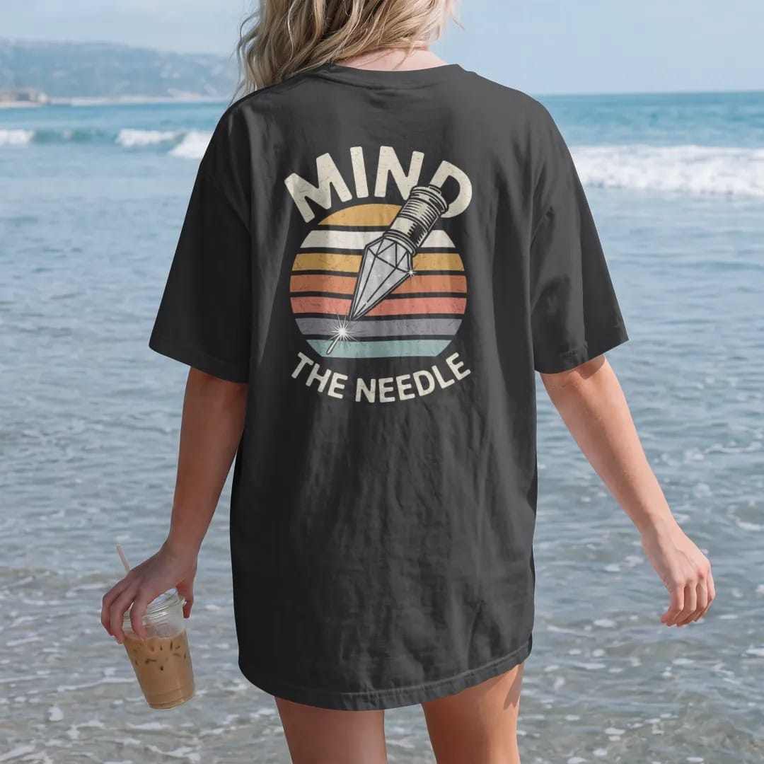 Woman on the beach in a Black tee, back view: a huge diamond stylus tip drawn like a jewel, with "MIND" and "THE NEEDLE" in chunky 70s lettering and small 70s caps, printed in faded, cracked cream, mustard, burnt orange and faded teal over faded…