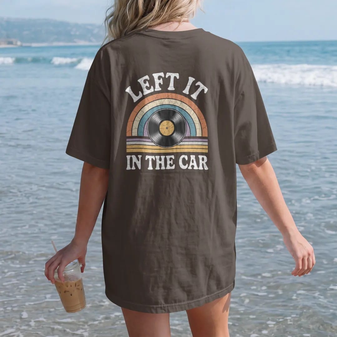 Woman on the beach in a Espresso tee, back view: a record melting over a car steering wheel under a hot sun, with "LEFT IT" and "IN THE CAR" in chunky 70s lettering and small 70s caps, printed in faded, cracked cream, mustard, burnt orange and…