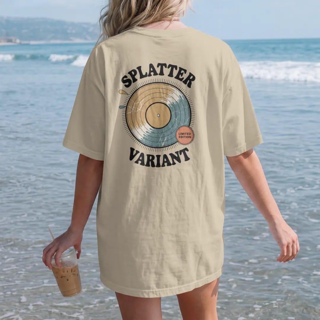 Woman on the beach in a Sandstone tee, back view: a paint-splatter colored record with a limited-edition sticker, with "SPLATTER" and "VARIANT" in chunky 70s lettering and small 70s caps, printed in faded, cracked cream, mustard, burnt orange and…