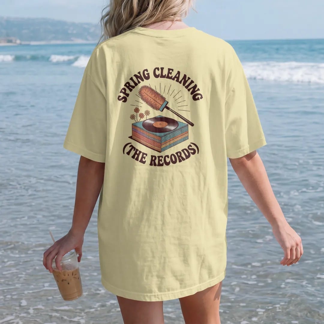 Woman on the beach in a Butter tee, back view: a feather duster and record brush over a stack sprouting spring flowers, with "SPRING CLEANING" and "(THE RECORDS)" in chunky 70s lettering and small 70s caps, printed in faded, cracked brown, burnt…