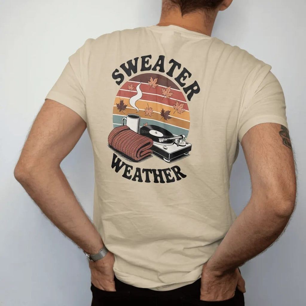 Man wearing a Sandstone tee, seen from the back: a mug, knit sweater and turntable among falling leaves, with "SWEATER WEATHER" and "SLOW SPINS" in chunky 70s lettering and small 70s caps, printed in faded, cracked brown, burnt orange, faded teal…