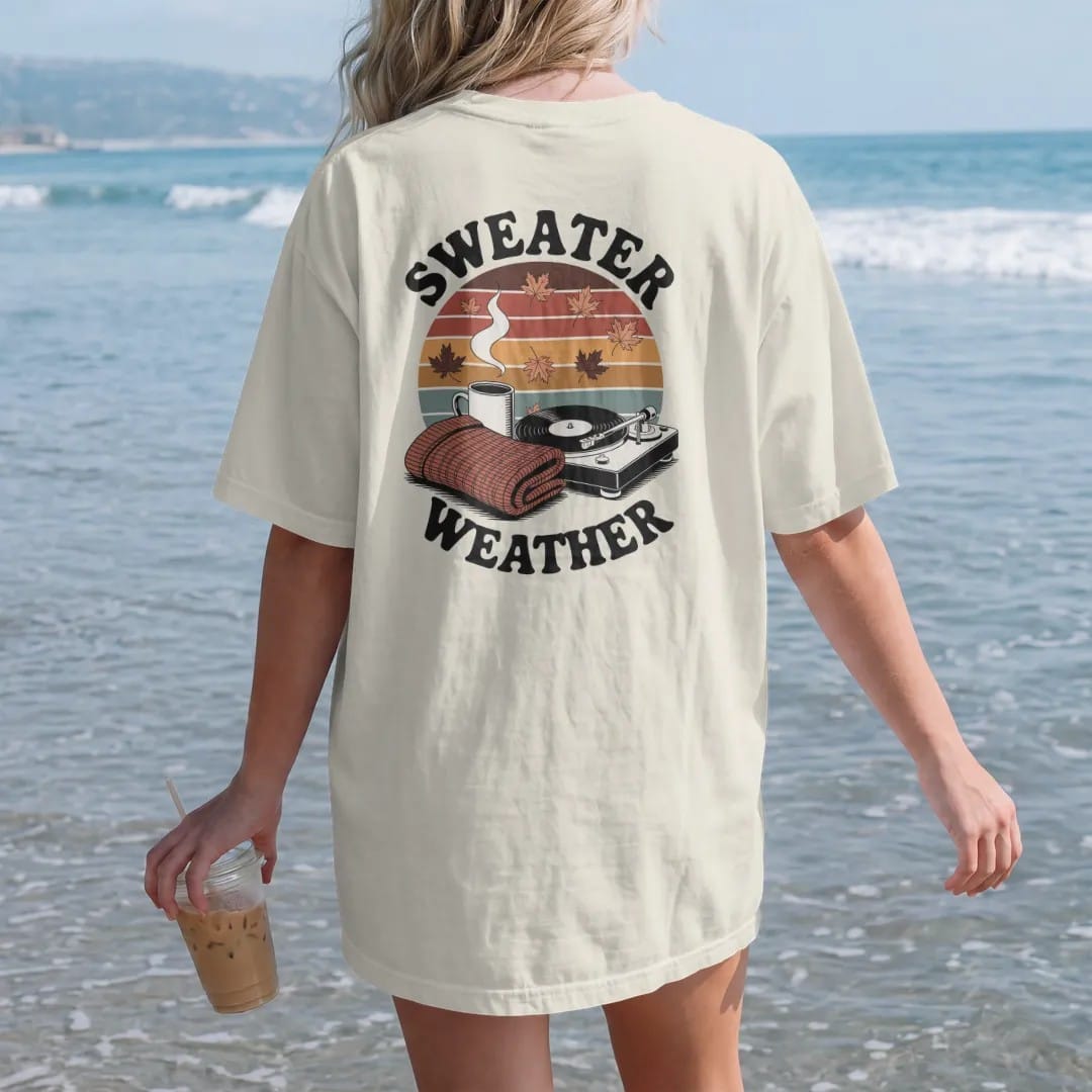 Woman on the beach in a Ivory tee, back view: a mug, knit sweater and turntable among falling leaves, with "SWEATER WEATHER" and "SLOW SPINS" in chunky 70s lettering and small 70s caps, printed in faded, cracked brown, burnt orange, faded teal and…