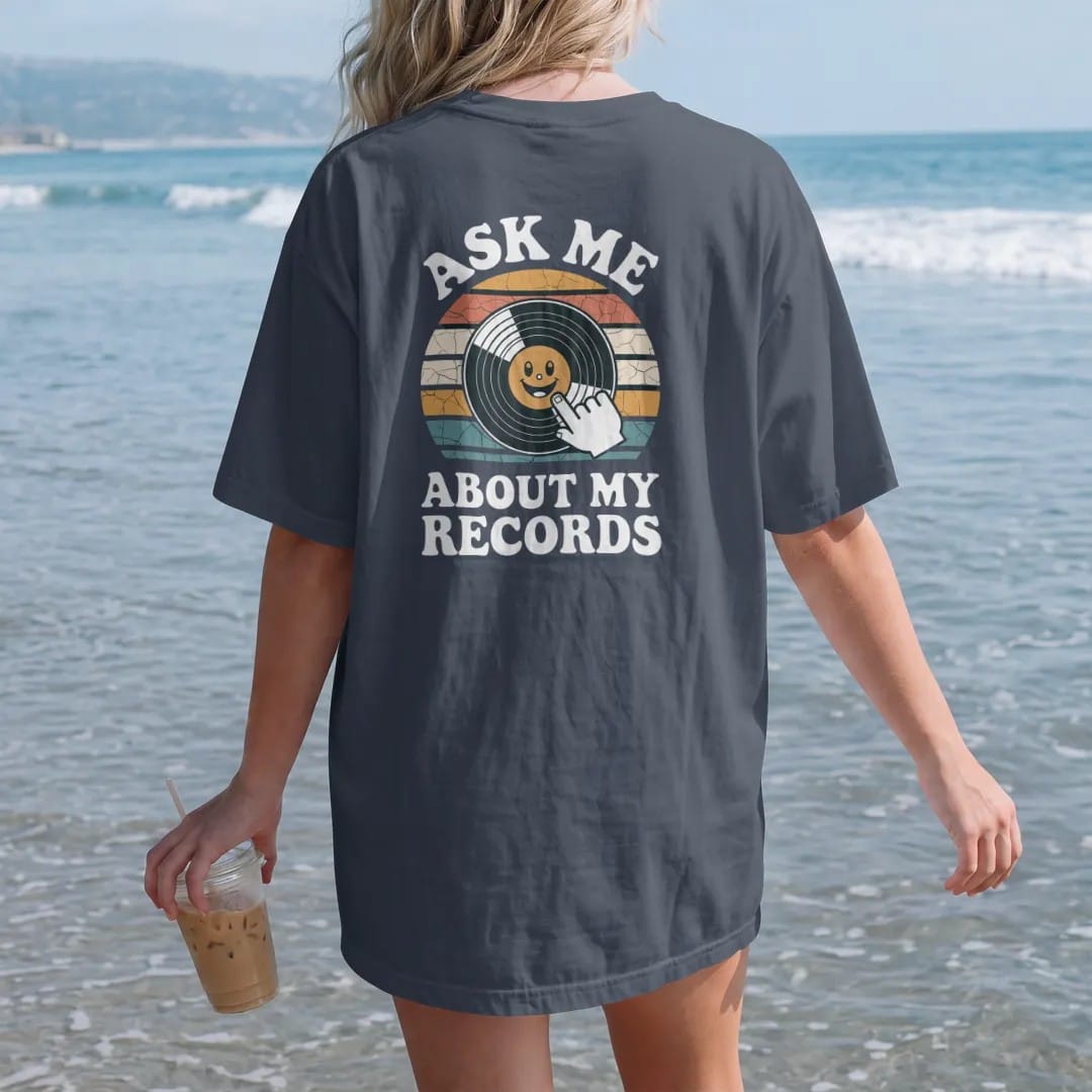 Woman on the beach in a Midnight tee, back view: a smiling record with a speech bubble and a pointing hand, with "ASK ME", "ABOUT MY" and "RECORDS" in chunky 70s lettering and small 70s caps, printed in faded, cracked cream, mustard, burnt orange…