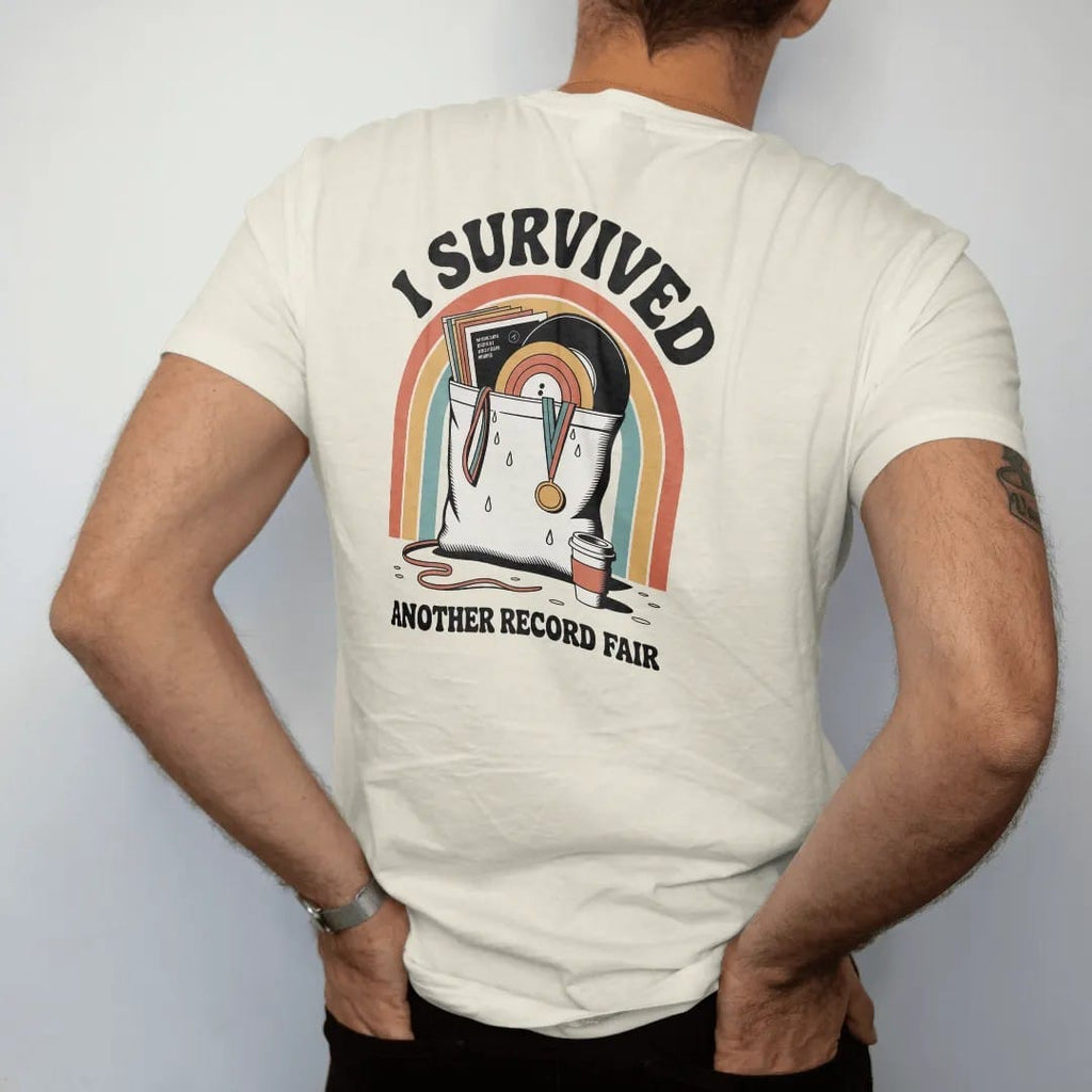 Man wearing a Ivory tee, seen from the back: an overstuffed tote bag of records with a medal ribbon, with "I SURVIVED" and "ANOTHER RECORD FAIR" in chunky 70s lettering and small 70s caps, printed in faded, cracked cream, mustard, burnt orange and…
