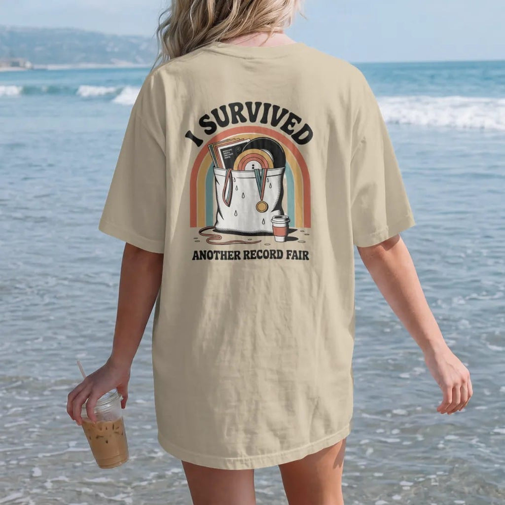 Woman on the beach in a Sandstone tee, back view: an overstuffed tote bag of records with a medal ribbon, with "I SURVIVED" and "ANOTHER RECORD FAIR" in chunky 70s lettering and small 70s caps, printed in faded, cracked cream, mustard, burnt orange…