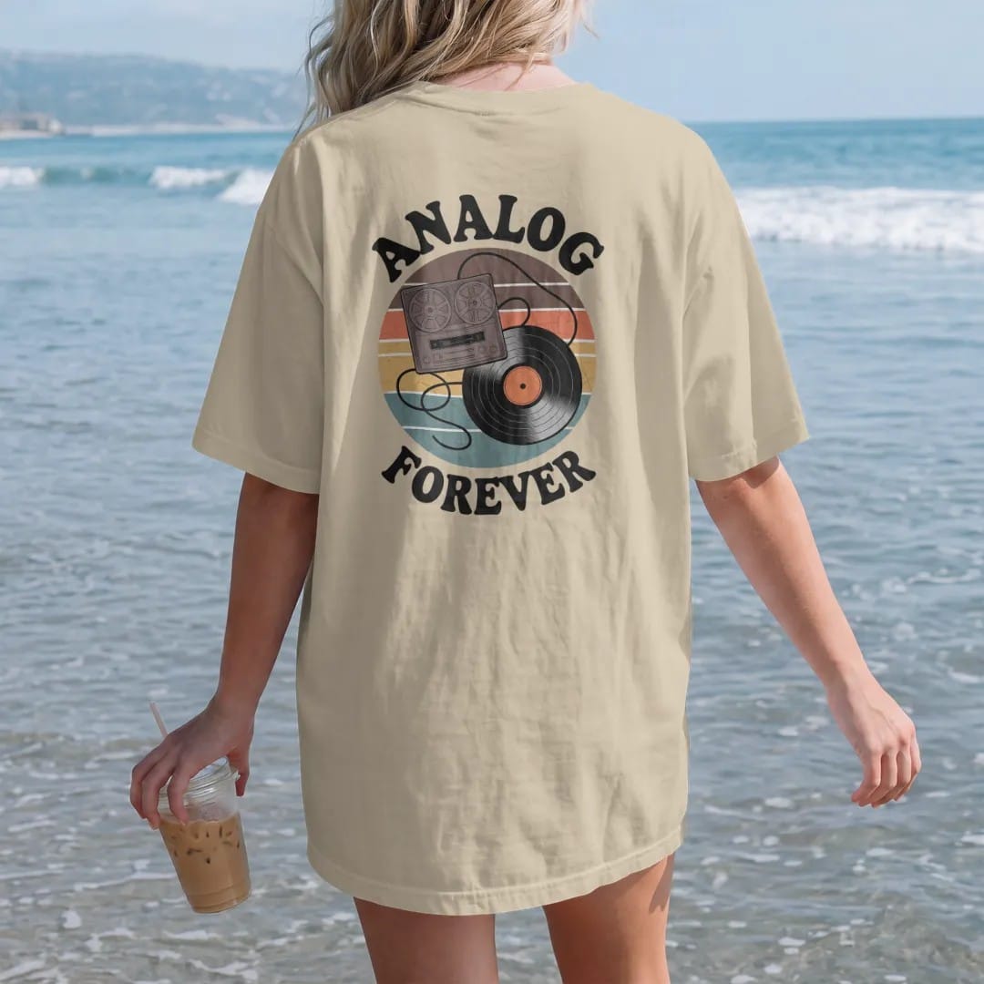 Woman on the beach in a Sandstone tee, back view: a reel-to-reel and a record joined by an infinity-loop tape ribbon, with "ANALOG" and "FOREVER" in chunky 70s lettering and small 70s caps, printed in faded, cracked brown, burnt orange, faded teal…