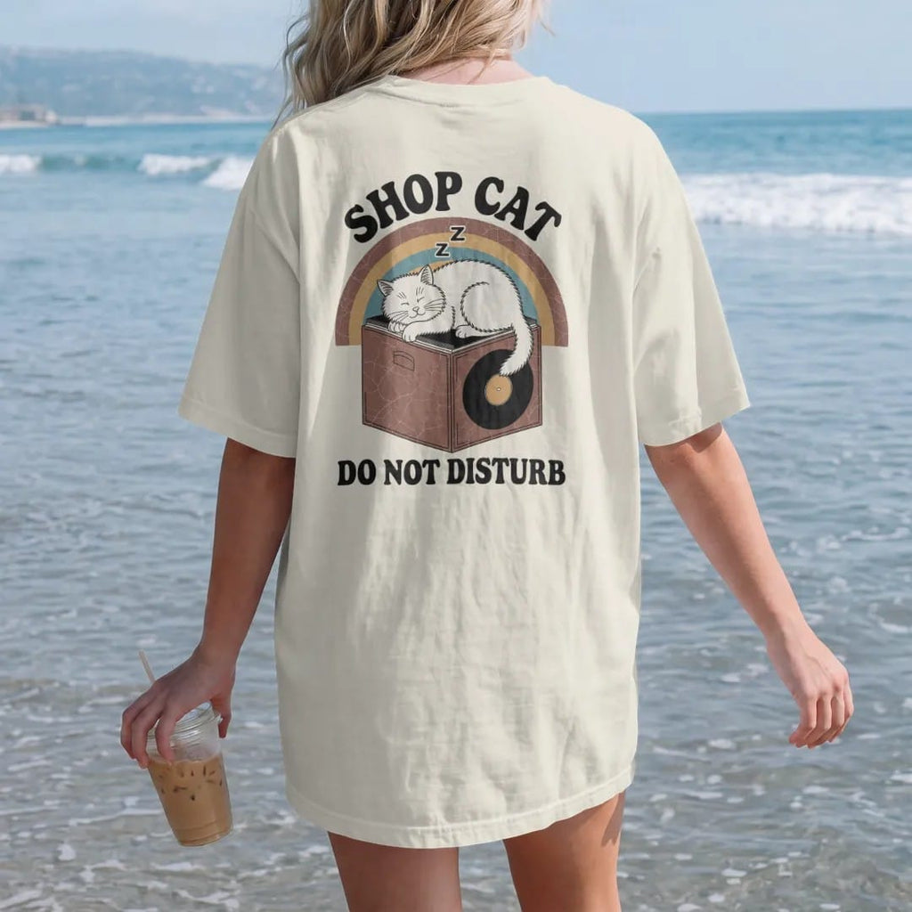 Woman on the beach in a Ivory tee, back view: a fluffy cat asleep on top of a record bin, with "SHOP CAT" and "DO NOT DISTURB" in chunky 70s lettering and small 70s caps, printed in faded, cracked brown, burnt orange, faded teal and mustard over a…