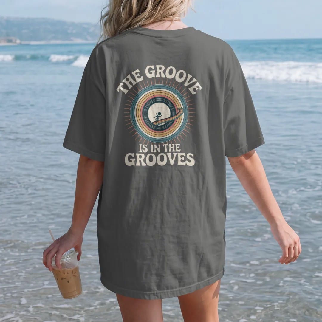 Woman on the beach in a Pepper tee, back view: macro record grooves as wavy lines with a tiny dancer surfing one, with "THE GROOVE" and "IS IN THE GROOVES" in chunky 70s lettering and small 70s caps, printed in faded, cracked cream, mustard, burnt…