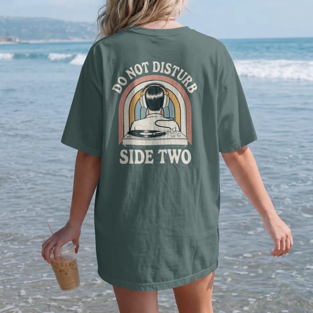 Woman on the beach in a Blue Spruce tee, back view: a person in over-ear headphones with a cord leading to a turntable, with "DO NOT DISTURB" and "SIDE TWO" in chunky 70s lettering and small 70s caps, printed in faded, cracked cream, mustard, burnt…