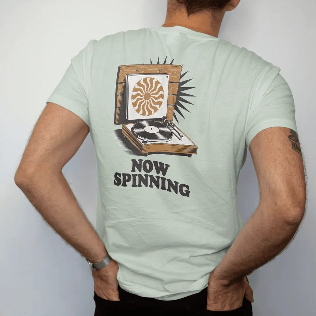 Man wearing a Bay tee, seen from the back: an album sleeve on a wall ledge above a spinning turntable, with "NOW SPINNING" in chunky 70s lettering, printed in faded, cracked brown, burnt orange, faded teal and mustard over a small sunburst.