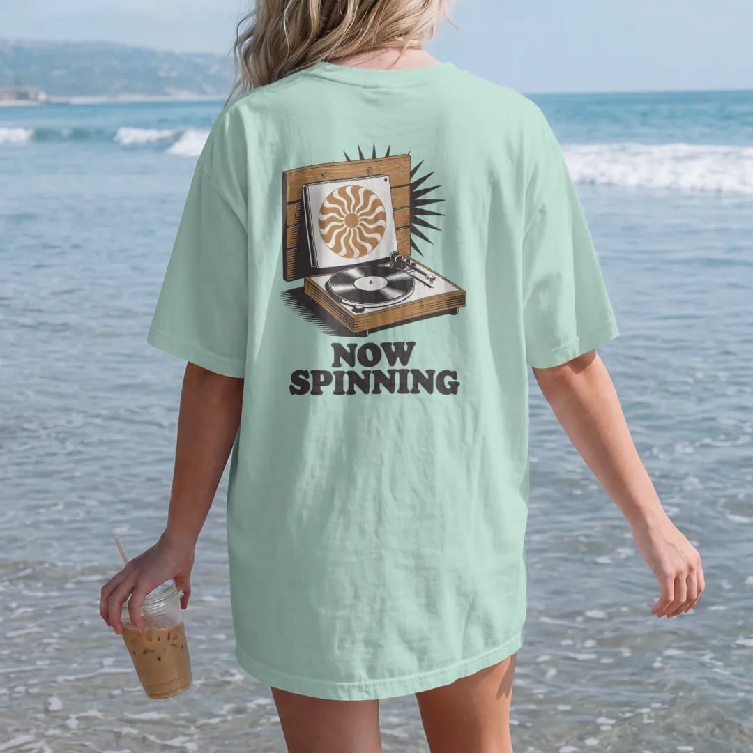 Woman on the beach in a Chalky Mint tee, back view: an album sleeve on a wall ledge above a spinning turntable, with "NOW SPINNING" in chunky 70s lettering, printed in faded, cracked brown, burnt orange, faded teal and mustard over a small sunburst.