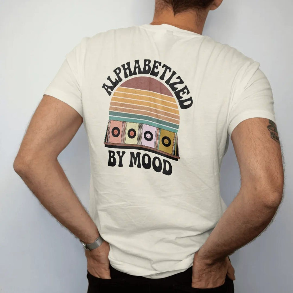Man wearing a Ivory tee, seen from the back: a shelf of record spines separated by mood-face divider cards, with "ALPHABETIZED" and "BY MOOD" in chunky 70s lettering and small 70s caps, printed in faded, cracked brown, burnt orange, faded teal and…