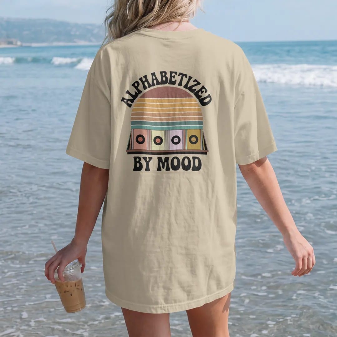 Woman on the beach in a Sandstone tee, back view: a shelf of record spines separated by mood-face divider cards, with "ALPHABETIZED" and "BY MOOD" in chunky 70s lettering and small 70s caps, printed in faded, cracked brown, burnt orange, faded teal…