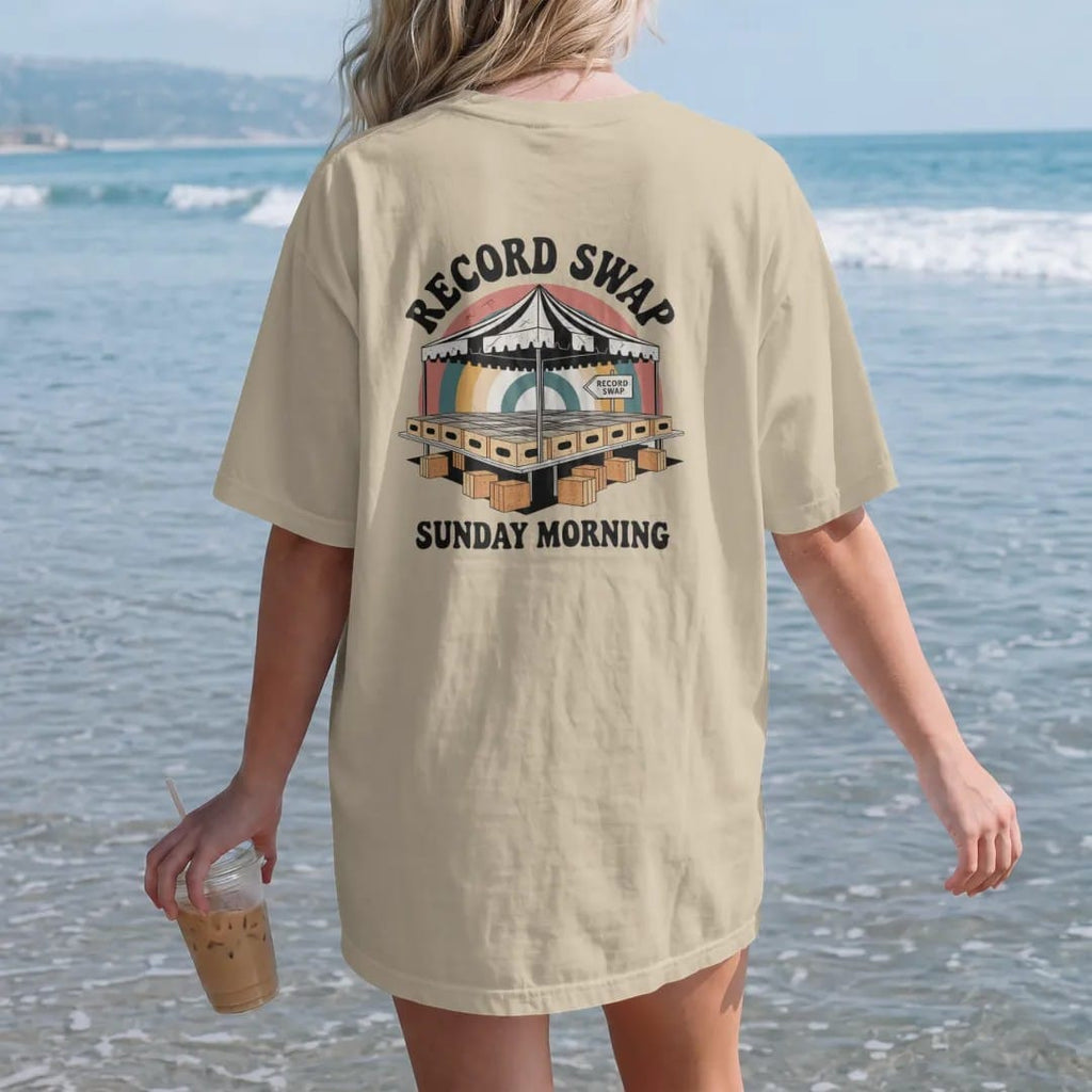 Woman on the beach in a Sandstone tee, back view: folding tables of record crates under a striped tent, with "RECORD SWAP" and "SUNDAY MORNING" in chunky 70s lettering and small 70s caps, printed in faded, cracked cream, mustard, burnt orange and…