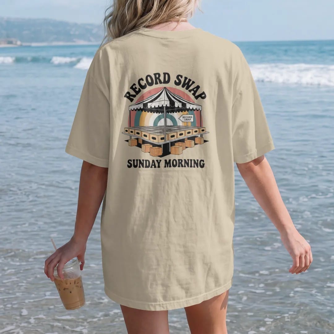 Woman on the beach in a Sandstone tee, back view: folding tables of record crates under a striped tent, with "RECORD SWAP" and "SUNDAY MORNING" in chunky 70s lettering and small 70s caps, printed in faded, cracked cream, mustard, burnt orange and…