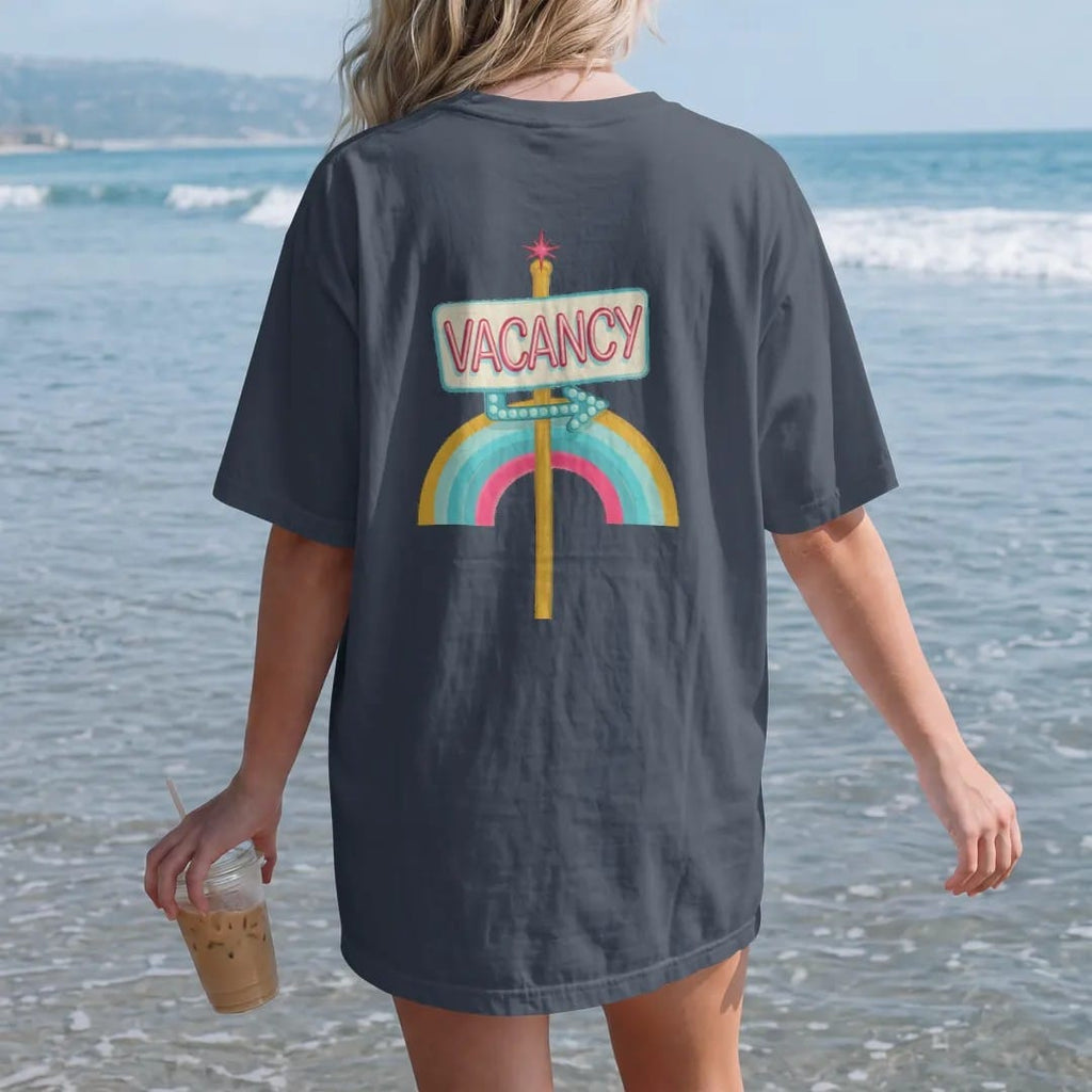 Woman on the beach in a Midnight tee, back view: a glowing neon vacancy sign on a pole with a bulb-dot arrow, with "VACANCY" in neon-tube script, printed in faded, cracked cream, mustard and turquoise with a neon pink accent over a faded rainbow arc.