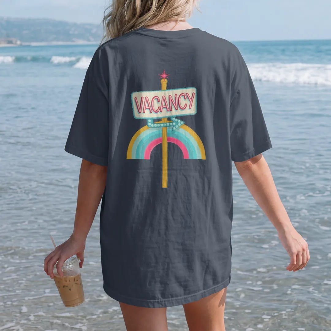 Woman on the beach in a Midnight tee, back view: a glowing neon vacancy sign on a pole with a bulb-dot arrow, with "VACANCY" in neon-tube script, printed in faded, cracked cream, mustard and turquoise with a neon pink accent over a faded rainbow arc.