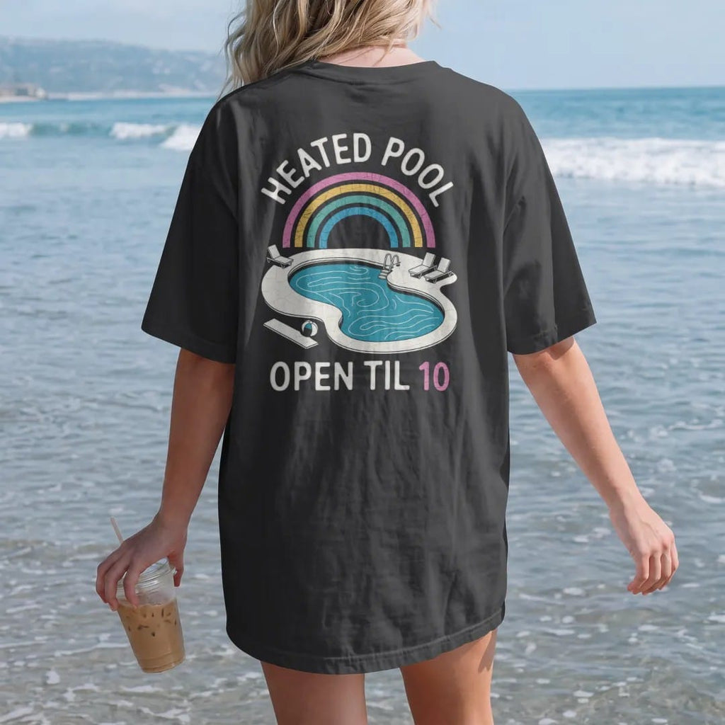 Woman on the beach in a Black tee, back view: a kidney-shaped motel pool from above with loungers and a diving board, with "HEATED POOL" and "OPEN TIL 10" in neon-tube script and small 70s caps, printed in faded, cracked cream, mustard and…