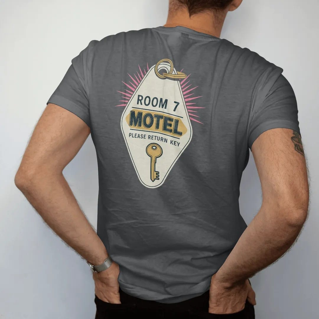 Man wearing a Graphite tee, seen from the back: an oversized diamond motel key fob with a brass key, with "ROOM 7" and "PLEASE RETURN KEY" in chunky 70s lettering and small 70s caps, printed in faded, cracked cream, mustard and turquoise with a…