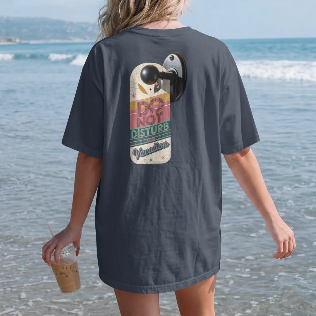Woman on the beach in a Midnight tee, back view: a retro door hanger with a sleeping moon on a motel doorknob, with "DO NOT DISTURB" and "ON VACATION" in chunky 70s lettering and brush script, printed in faded, cracked cream, mustard and turquoise…