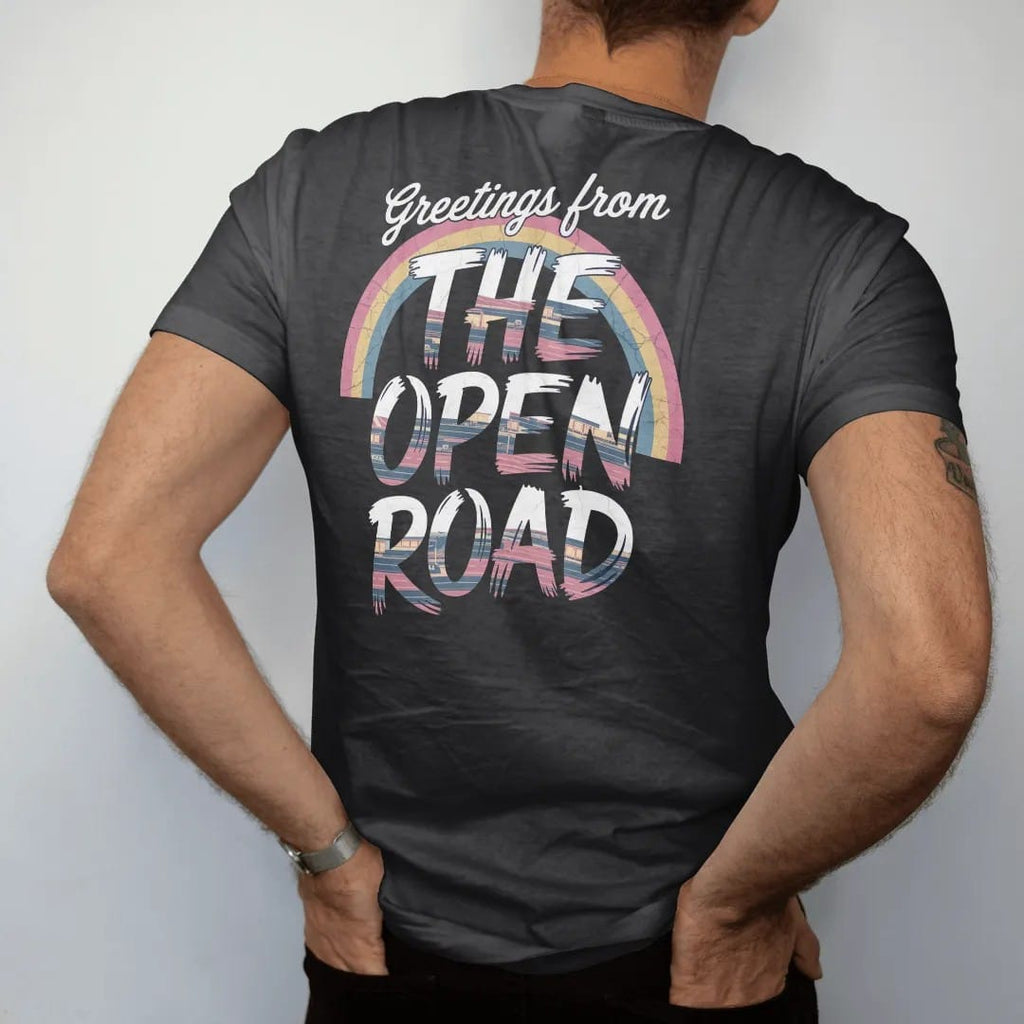 Man wearing a Black tee, seen from the back: a big-letter postcard with road-trip scenes inside chunky letters, with "GREETINGS FROM" and "THE OPEN ROAD" in chunky 70s lettering and brush script, printed in faded, cracked navy, rust red and dusty…