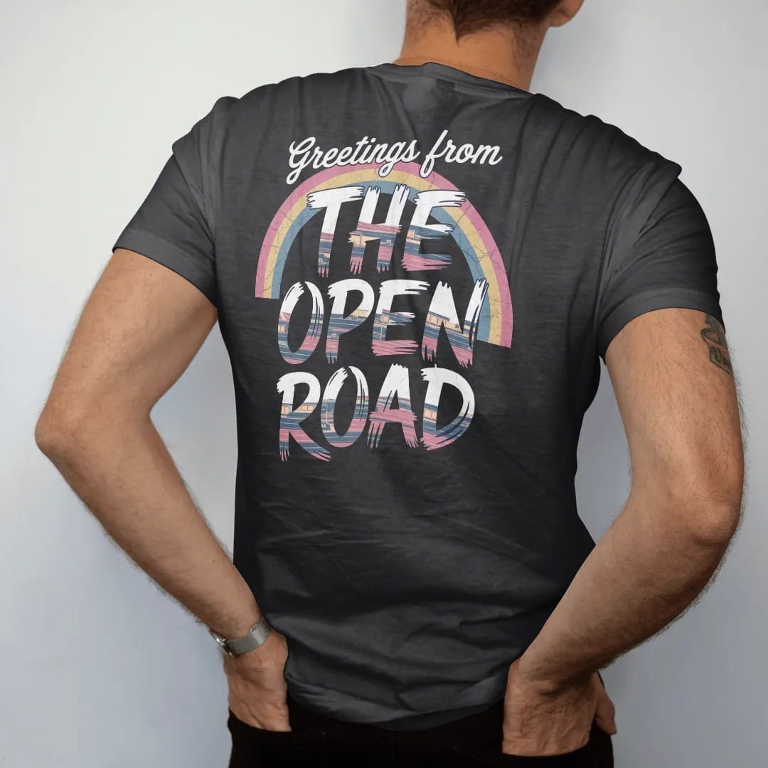Man wearing a Black tee, seen from the back: a big-letter postcard with road-trip scenes inside chunky letters, with "GREETINGS FROM" and "THE OPEN ROAD" in chunky 70s lettering and brush script, printed in faded, cracked navy, rust red and dusty…