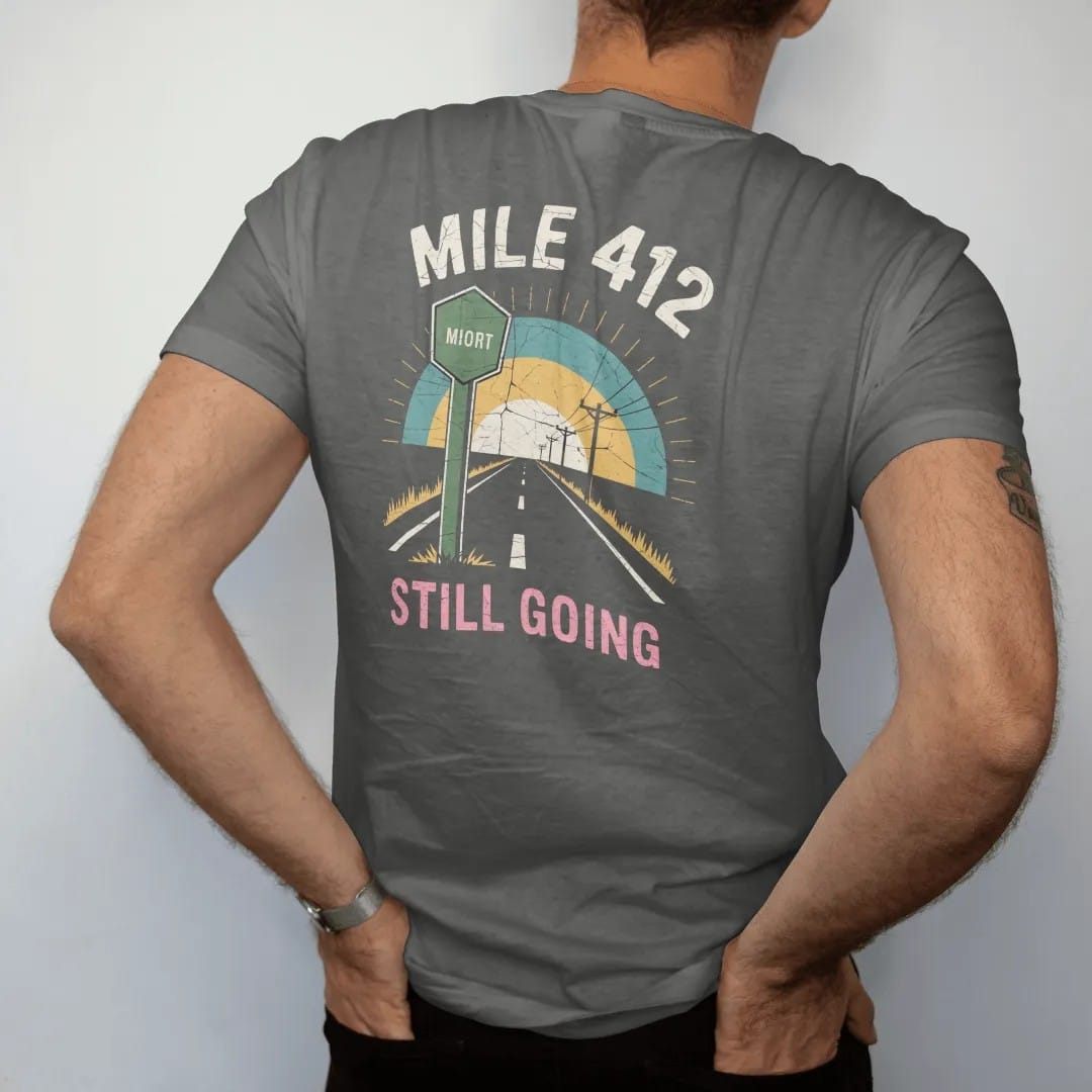Man wearing a Pepper tee, seen from the back: a highway mile marker beside a two-lane road at dusk, with "MILE 412" and "STILL GOING" in chunky 70s lettering and small 70s caps, printed in faded, cracked cream, mustard and turquoise with a neon…
