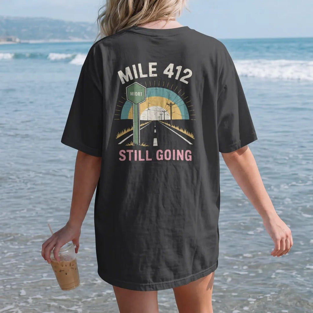 Woman on the beach in a Black tee, back view: a highway mile marker beside a two-lane road at dusk, with "MILE 412" and "STILL GOING" in chunky 70s lettering and small 70s caps, printed in faded, cracked cream, mustard and turquoise with a neon…