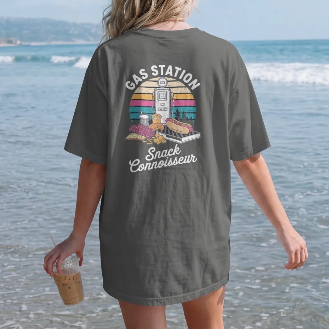 Woman on the beach in a Pepper tee, back view: a pile of road-trip snacks in a gourmet crest frame, with "GAS STATION" and "SNACK CONNOISSEUR" in chunky 70s lettering and brush script, printed in faded, cracked cream, mustard and turquoise with a…