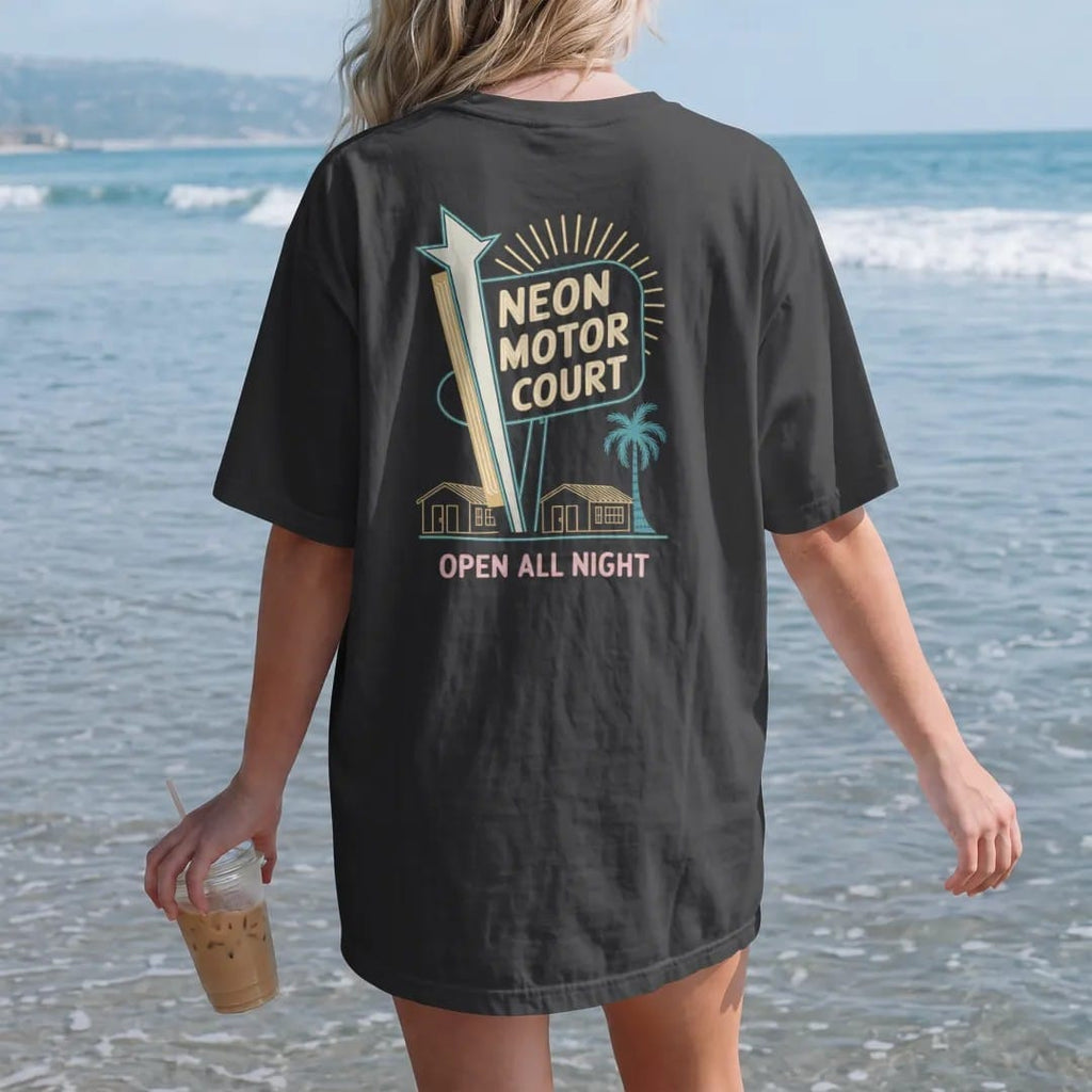 Woman on the beach in a Black tee, back view: a tall neon motel sign with a starburst over little bungalows, with "NEON MOTOR COURT" and "OPEN ALL NIGHT" in neon-tube script and small 70s caps, printed in faded, cracked cream, mustard and turquoise…